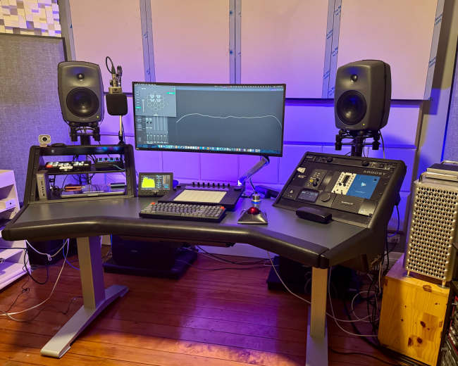The image depicts a music production studio featuring a desk with a computer monitor, audio interface, speakers, and various audio equipment.