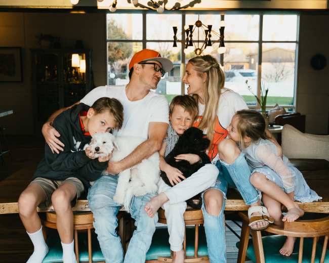A family sits together on a wooden table in a brightly lit room, each holding a dog, with children smiling beside their parents.