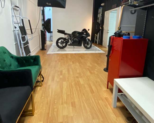 A spacious studio with a motorcycle displayed on a white platform, surrounded by studio lighting and various props.