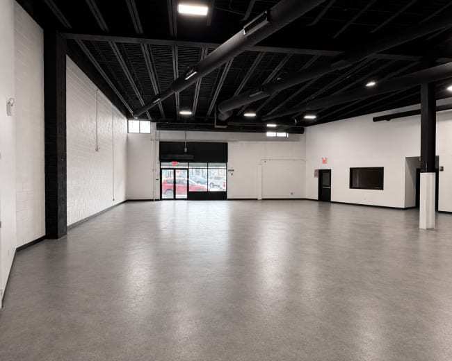 The image shows a large, empty room with gray flooring, black beams overhead, and a doorway leading outside.