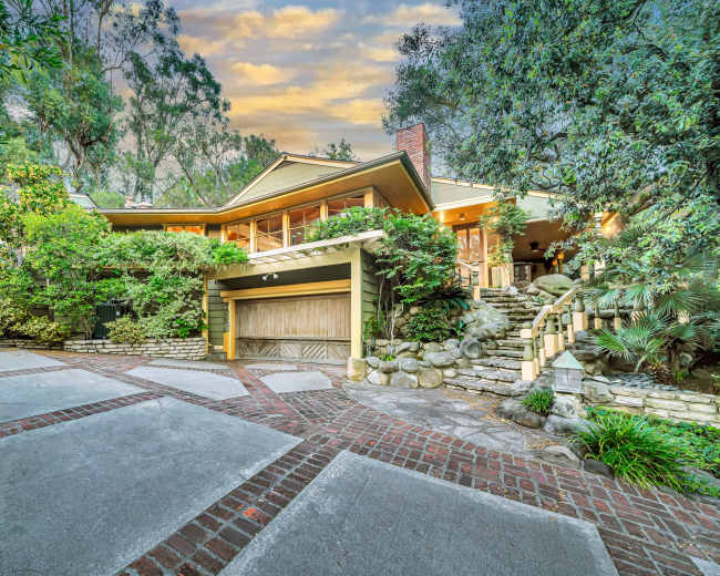 A multi-level house with a stone path and lush greenery is surrounded by trees in a tranquil setting.