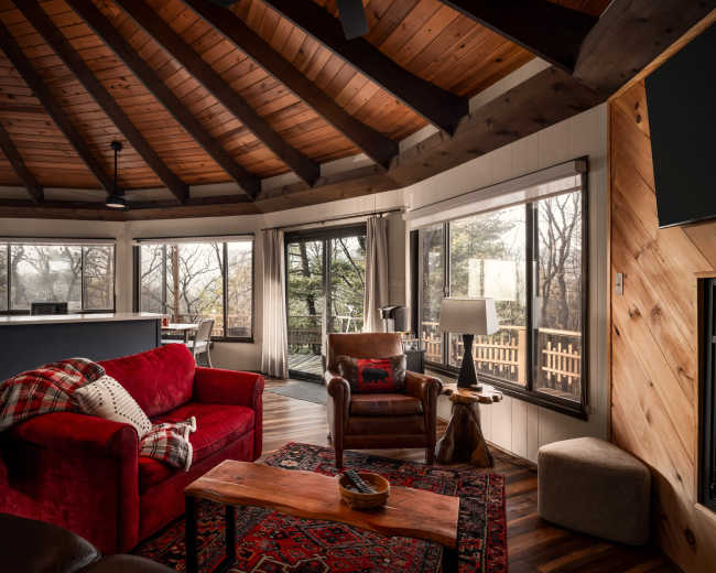 The image shows a cozy living room with a vaulted wooden ceiling, large windows, a red sofa, and a wooden coffee table.