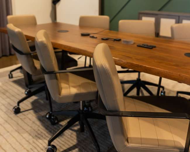 A long wooden conference table with several swivel chairs is positioned in a meeting room featuring green paneled walls.