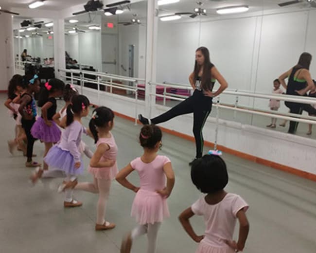 A dance instructor demonstrates a move to a group of young students in a mirrored studio.
