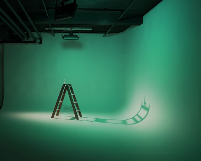 A wooden ladder stands alone in an illuminated studio space with a green backdrop.
