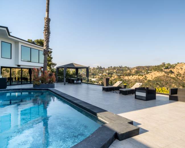 The image shows a modern outdoor patio with a swimming pool, lounge chairs, and a gazebo overlooking a hilly landscape.