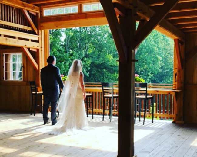 A bride and groom walk together on a wooden floor inside a cabin with large windows overlooking a forested area.