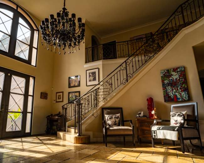 The image shows a well-lit entrance hall with a chandelier, a staircase, and stylish chairs featuring decorative face designs.