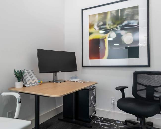 The image shows a minimalist office space with a wooden desk, a monitor, a potted plant, and two office chairs, all against a wall featuring a large framed abstract photo.