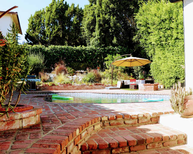 The image shows a landscaped backyard featuring a swimming pool surrounded by brick paving, lounge furniture, and an umbrella.