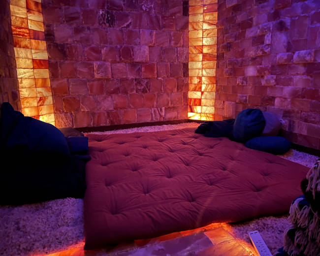 The image shows a dimly lit room with salt brick walls, featuring a red mat on the floor surrounded by pillows.