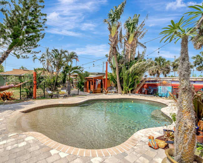 The image shows a landscaped backyard featuring a swimming pool surrounded by palm trees, a hammock, and a wooden structure.