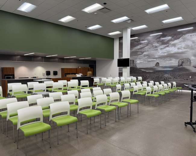 The image shows a modern indoor space with arranged rows of green and white chairs facing a counter area, complemented by a large wall mural of a landscape.