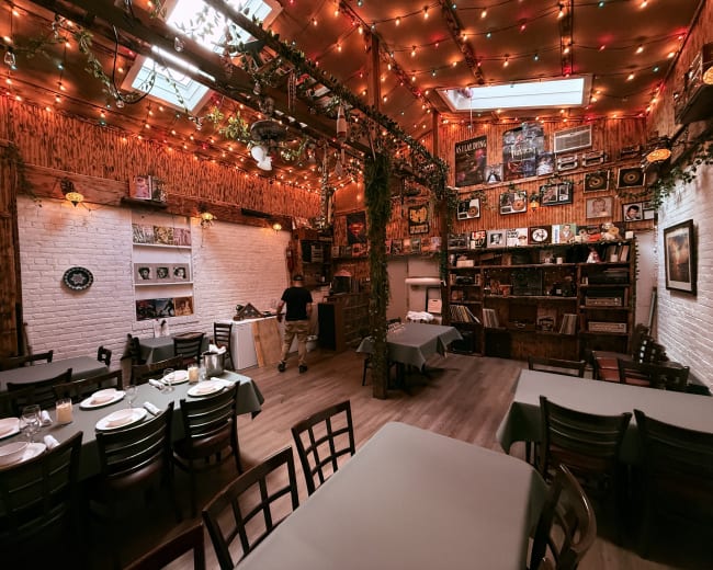 The image shows a warmly decorated restaurant interior with tables covered in green cloths, a wooden shelving unit filled with various items, and soft lighting from string lights.