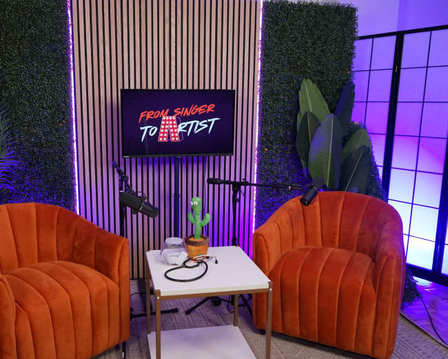 A set featuring two orange chairs, a small table with a plant and microphone cables, and a backdrop displaying the phrase "From Singer to Artist."