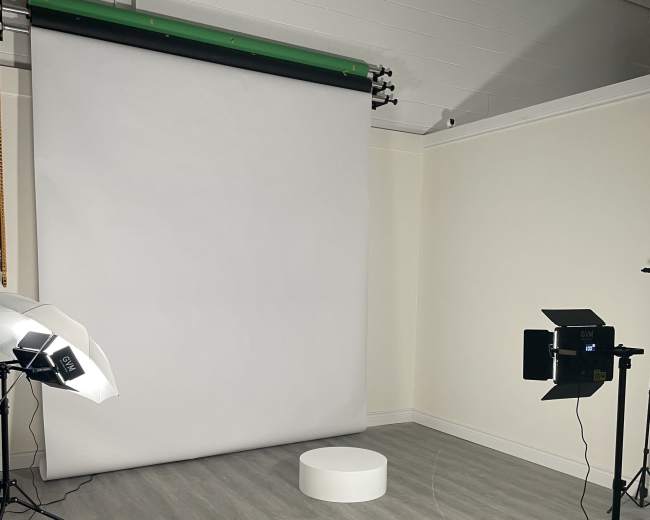The image shows a photography studio with a white backdrop, a circular platform in the center, and lighting equipment on either side.