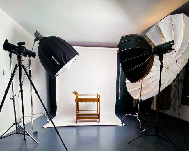 The image shows a photography studio with two large light sources positioned on either side of a wooden cart set against a white backdrop.