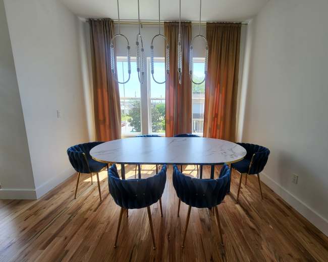 A modern dining area features a marble-topped oval table surrounded by blue velvet chairs and large windows adorned with gradient curtains.