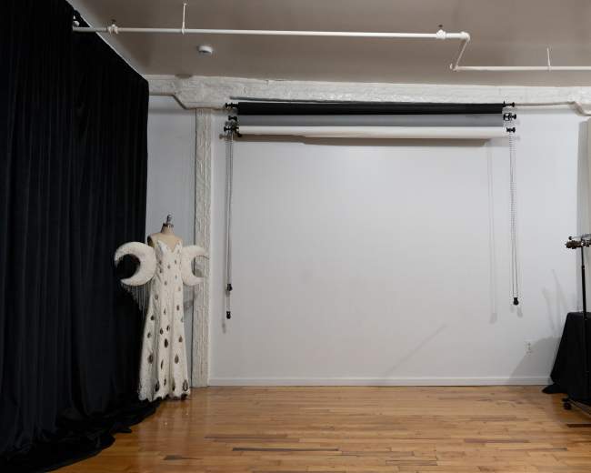 The image shows a studio space with a blank wall, a garment on a mannequin, and a backdrop support system on the ceiling, alongside a table with color rolls in the background.
