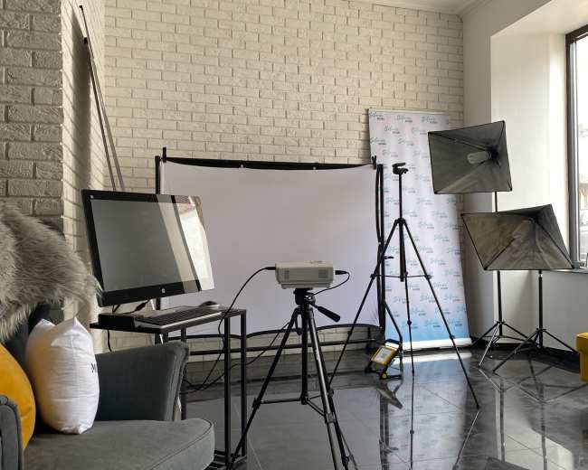 A room with a gray couch, a projector on a tripod, and two lighting setups in front of a white backdrop.