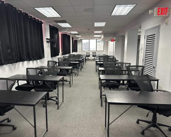 The image depicts a classroom or office space arranged with numerous black tables and chairs, evenly spaced in rows with large windows covered by black curtains.