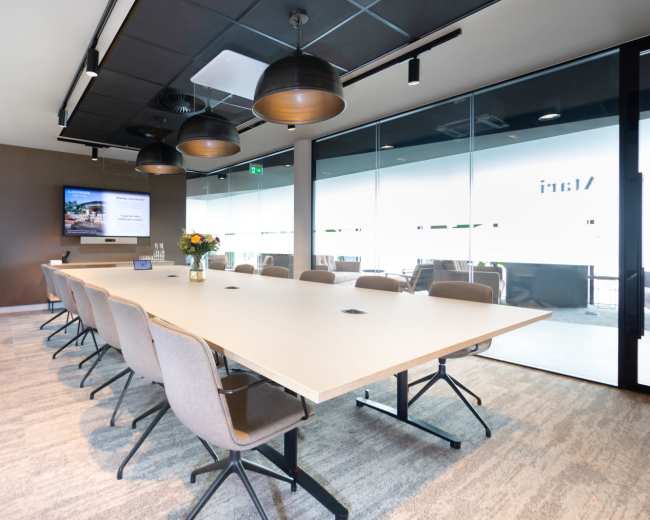 A modern conference room features a long table surrounded by gray chairs, with a large screen on the wall and windows displaying an outdoor view.