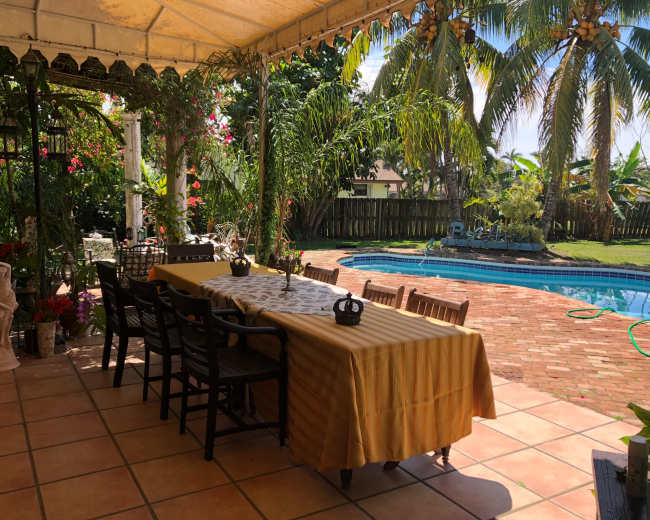 A large outdoor dining area with a long table under a canopy, surrounded by greenery and adjacent to a swimming pool.