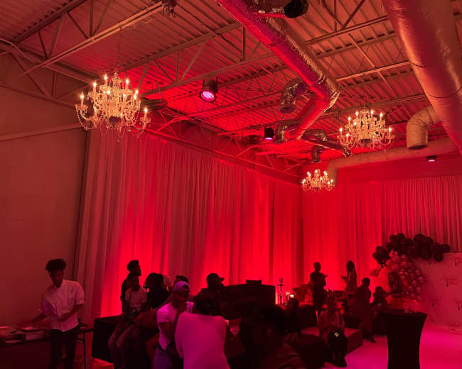 An indoor event space illuminated in red light, featuring chandeliers and a variety of guests gathered around tables.