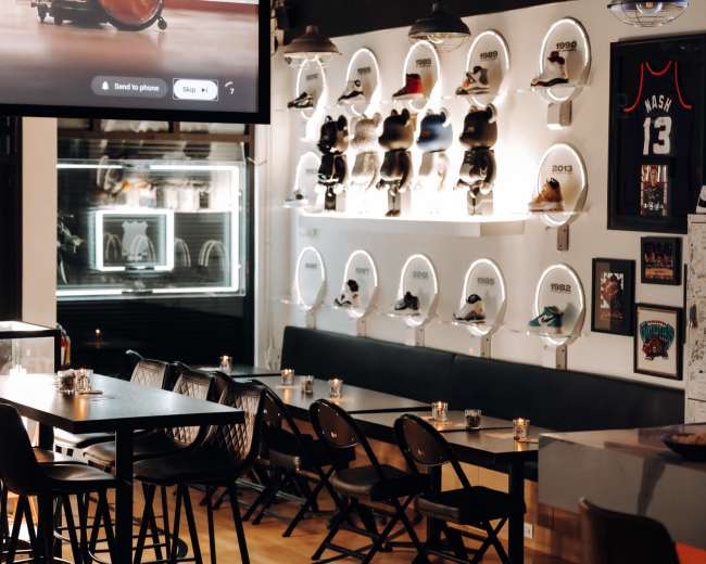 The image shows a modern cafe interior with a long table, black chairs, and a wall display featuring sneakers in clear cases, complemented by a large screen showing a video.