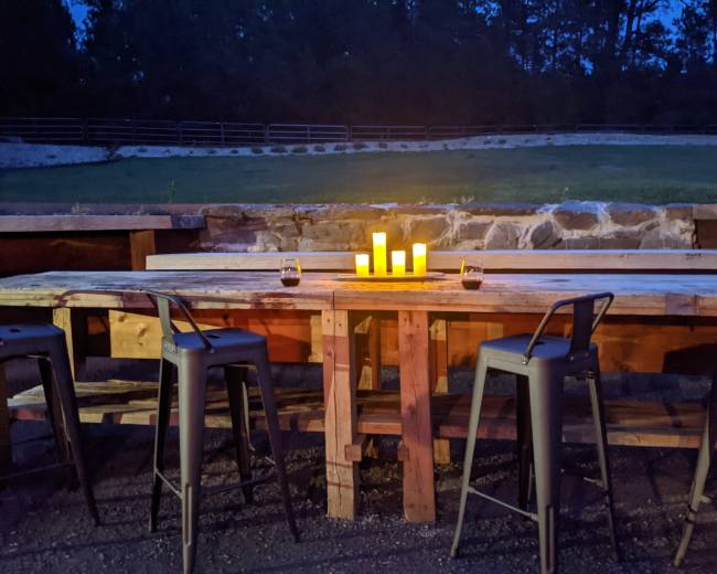 A wooden bar table with high stools is illuminated by candles in a dimly lit outdoor setting.