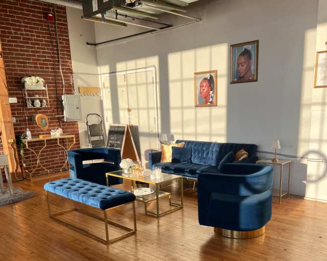 The image shows a well-lit interior space featuring a blue velvet sofa, matching benches, and framed portraits on the wall, all set against hardwood flooring.