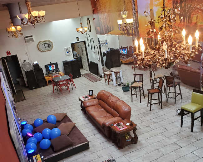 The image shows a spacious interior room with a brown leather sofa, several chairs, a DJ setup, and blue balloons scattered on the floor.