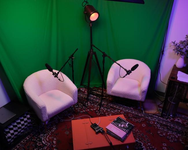 The image shows a recording setup featuring two white chairs with microphones, a green backdrop, a tripod light, and a table with audio equipment.