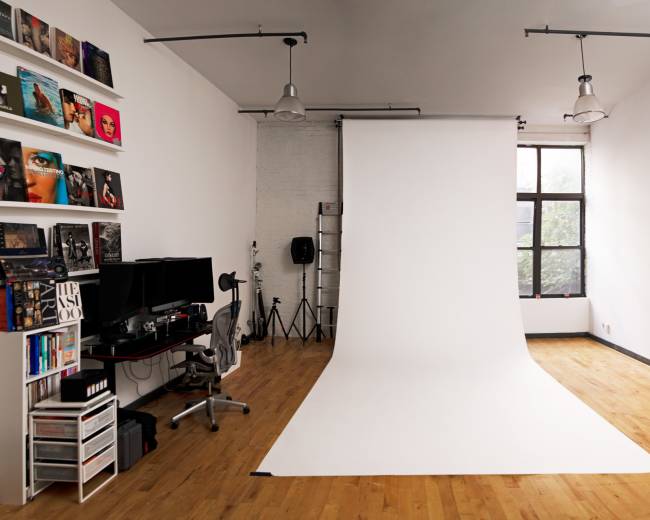 A photography studio features a white backdrop, wooden flooring, and equipment arranged near a large window.