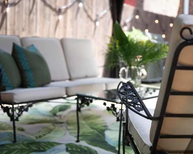 The image shows a patio setting with a metal-framed sofa and a glass coffee table on a leaf-patterned rug, surrounded by plants and string lights in the background.