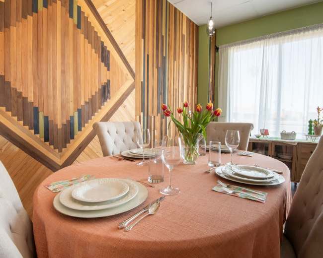 The image shows a dining table set with plates, glasses, and a vase of tulips, surrounded by upholstered chairs and a wooden accent wall.