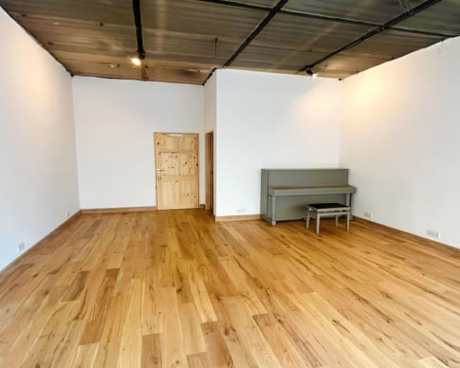 The image shows a spacious, empty room with hardwood floors, white walls, and a gray piano positioned against one wall.
