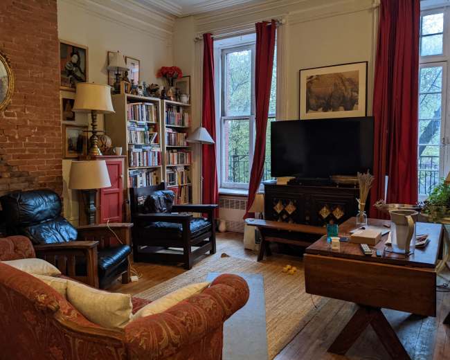 The living room features a mix of seating, a large bookshelf, a television, and a window with red curtains, set against a backdrop of exposed brick walls.