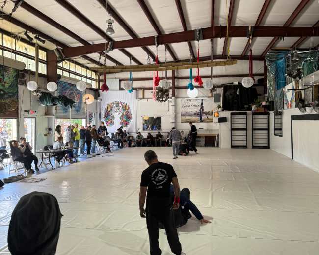 The image shows a spacious indoor training area with a white mat, people engaged in activities, and colorful decorations hanging from the ceiling.