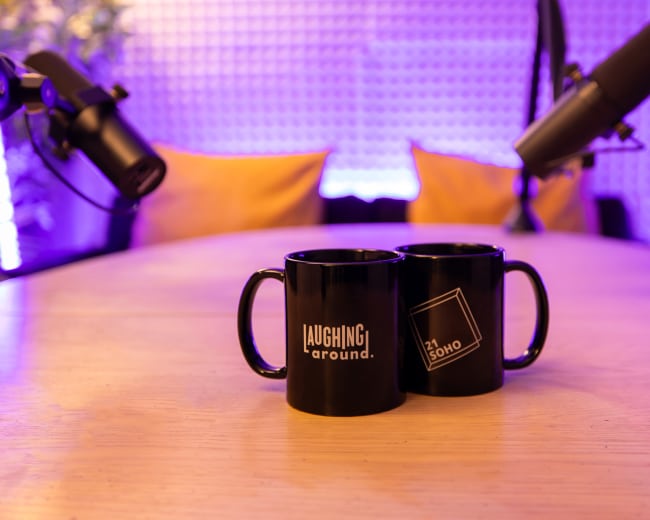 Two black mugs with distinct logos sit on a wooden table between two microphones in a studio setting.