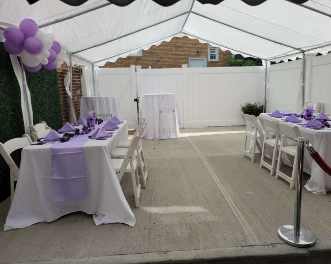 A tented outdoor space features tables set with lavender tablecloths and decorations, arranged for an event with a white fence in the background.