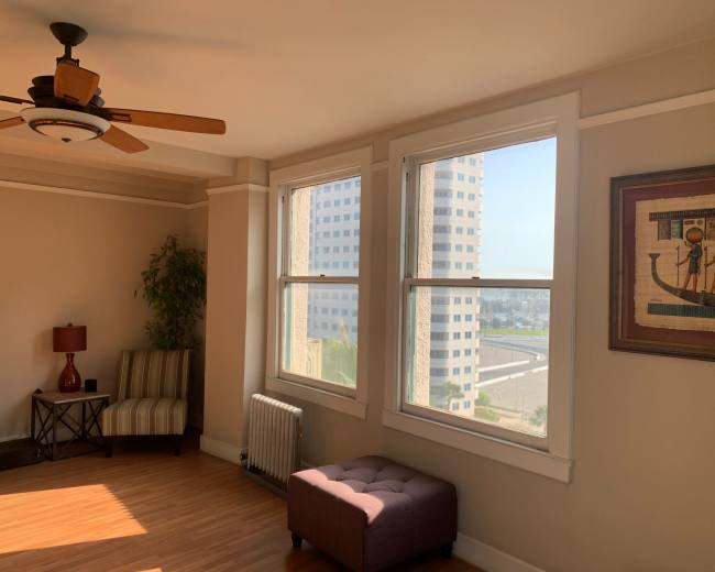 A bright living room with large windows, a ceiling fan, and a small seating area featuring a striped chair and a purple ottoman.