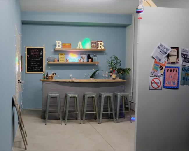 The image shows a bar area with a countertop, high stools, colorful neon lights, and a chalkboard menu in a light blue room.