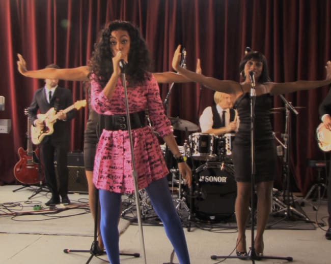 A female singer performs on stage with backup vocalists and a band in a studio setting.