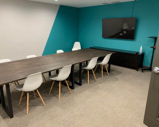 The image shows a conference room with a long wooden table surrounded by white chairs, a television mounted on the wall, and a side cabinet.