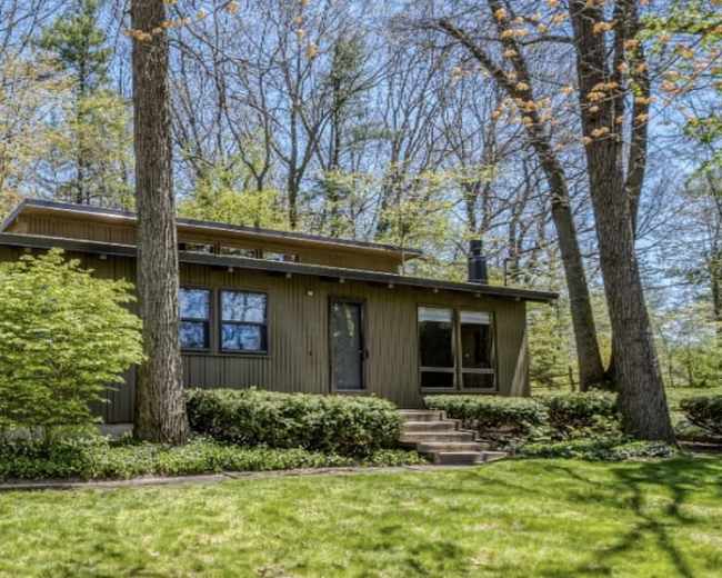 A single-story wooden house with large windows is surrounded by lush green grass and trees.