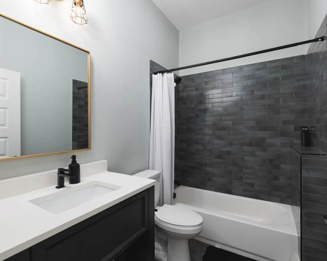 The image shows a modern bathroom featuring a black vanity with a white sink, a large mirror, a bathtub with a gray tiled wall, and a white shower curtain.