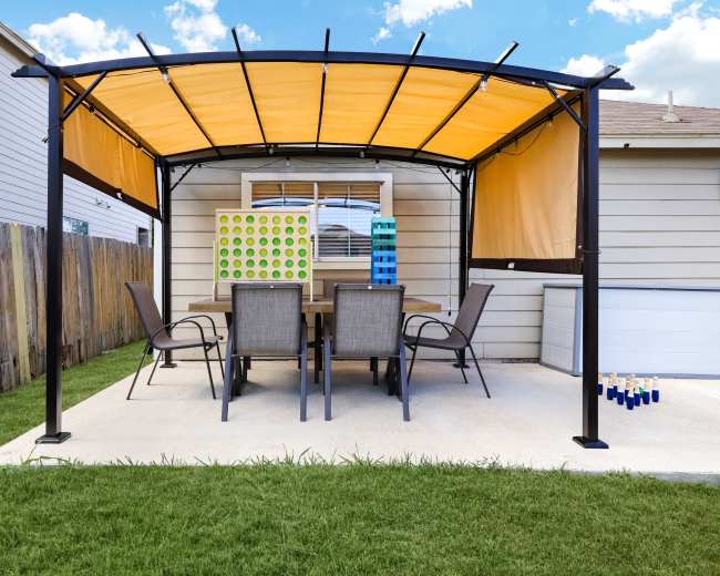 A patio with an orange canopy covers a table and six chairs in a backyard, with a grass lawn and a nearby shed.