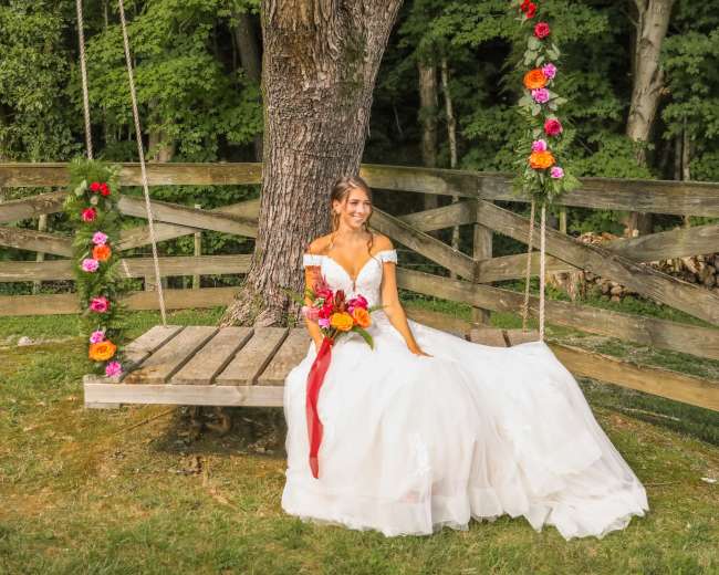 A bride in a flowing white dress sits on a wooden swing decorated with flowers, beneath a large tree in a grassy area.