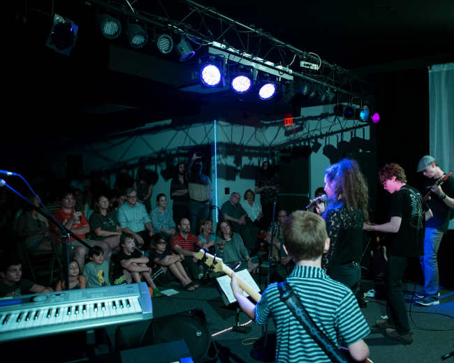 A group of young musicians performs on stage in front of an audience at a small venue.